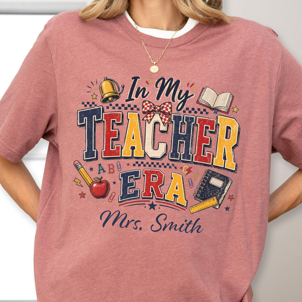 May include: A mauve short-sleeve t-shirt featuring a graphic with the text In My Teacher Era and Mrs. Smith. The design includes a gold bell, a red apple, a checkered bow, an open book, a spiral notebook, a pencil, and a ruler in red, blue, and yellow.