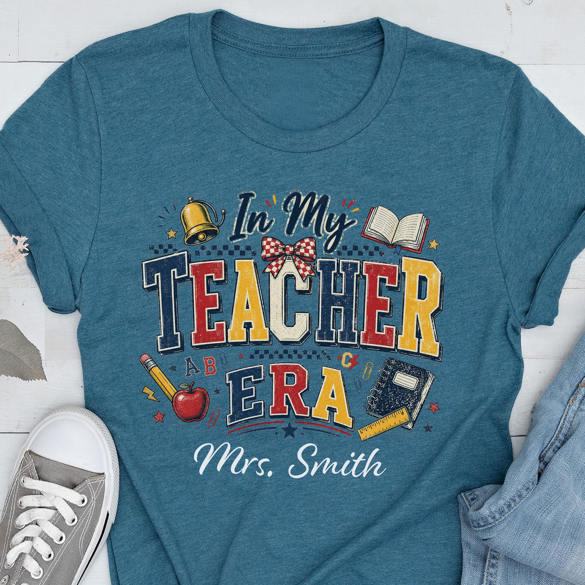 May include: A teal blue t-shirt featuring a graphic with the text In My Teacher Era and Mrs. Smith. The design includes a gold bell, an open book, a red apple, a pencil, and a notebook, all arranged in a colorful, school-themed composition.