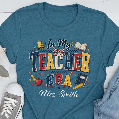 May include: A teal blue t-shirt featuring a graphic with the text In My Teacher Era and Mrs. Smith. The design includes a gold bell, an open book, a red apple, a pencil, and a notebook, all arranged in a colorful, school-themed composition.
