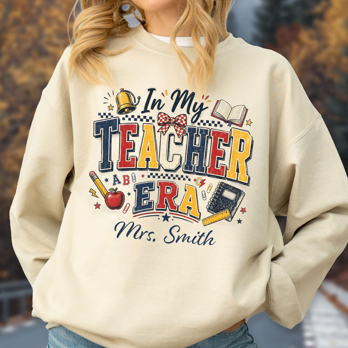 May include: A beige crewneck sweatshirt featuring a graphic print with the text In My TEACHER ERA and Mrs. Smith. The design includes illustrations of a gold bell, a red apple, a pencil, a notebook, a ruler, and a red and white checkered bow.