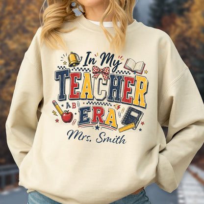 May include: A beige crewneck sweatshirt featuring a graphic print with the text In My TEACHER ERA and Mrs. Smith. The design includes illustrations of a gold bell, a red apple, a pencil, a notebook, a ruler, and a red and white checkered bow.
