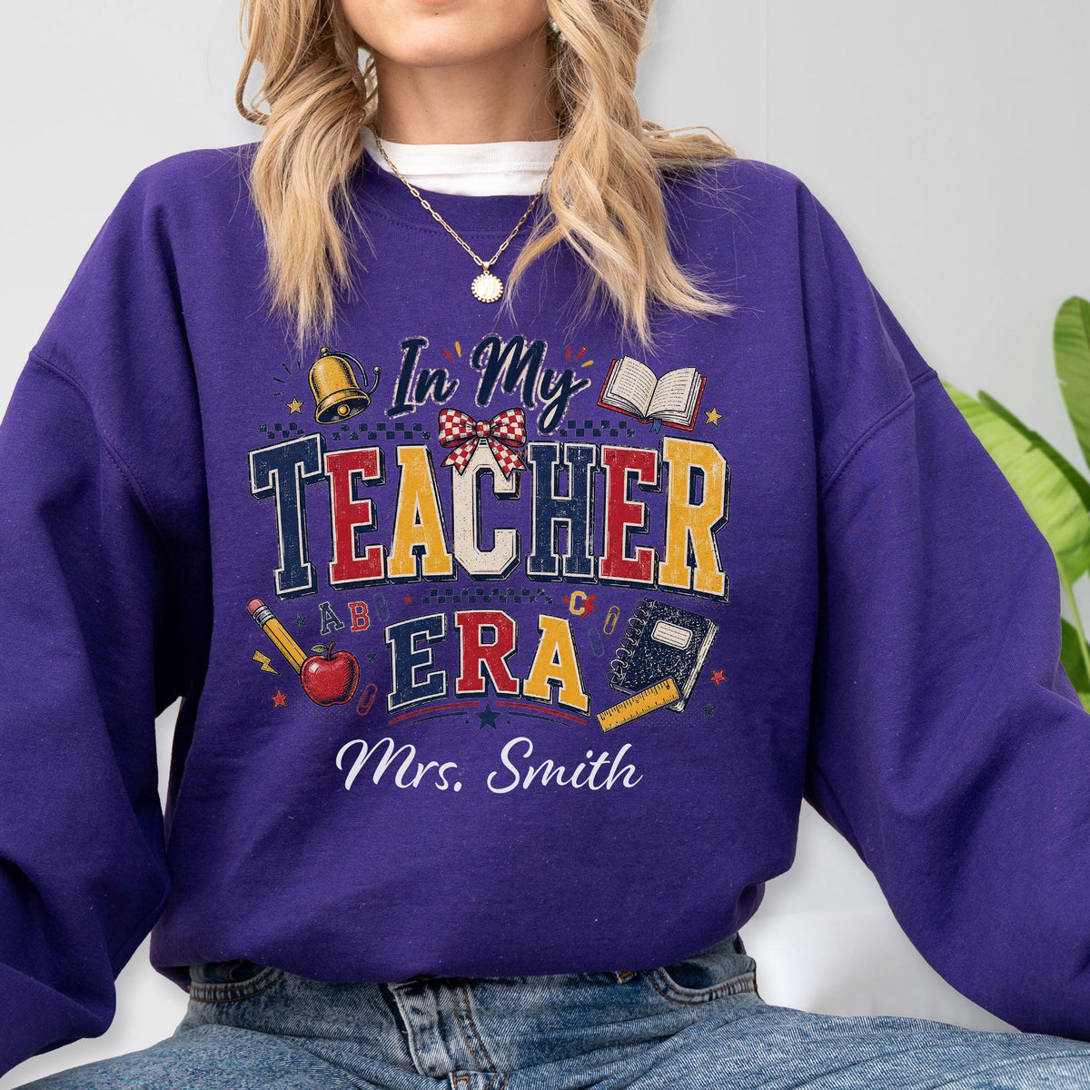 May include: A purple long-sleeve sweatshirt featuring a graphic design with the text In My Teacher Era and Mrs. Smith. The design includes illustrations of a gold bell, an open book, a red apple, a yellow pencil, and a blue notebook with a ruler.