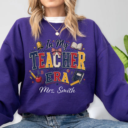 May include: A purple long-sleeve sweatshirt featuring a graphic design with the text In My Teacher Era and Mrs. Smith. The design includes illustrations of a gold bell, an open book, a red apple, a yellow pencil, and a blue notebook with a ruler.