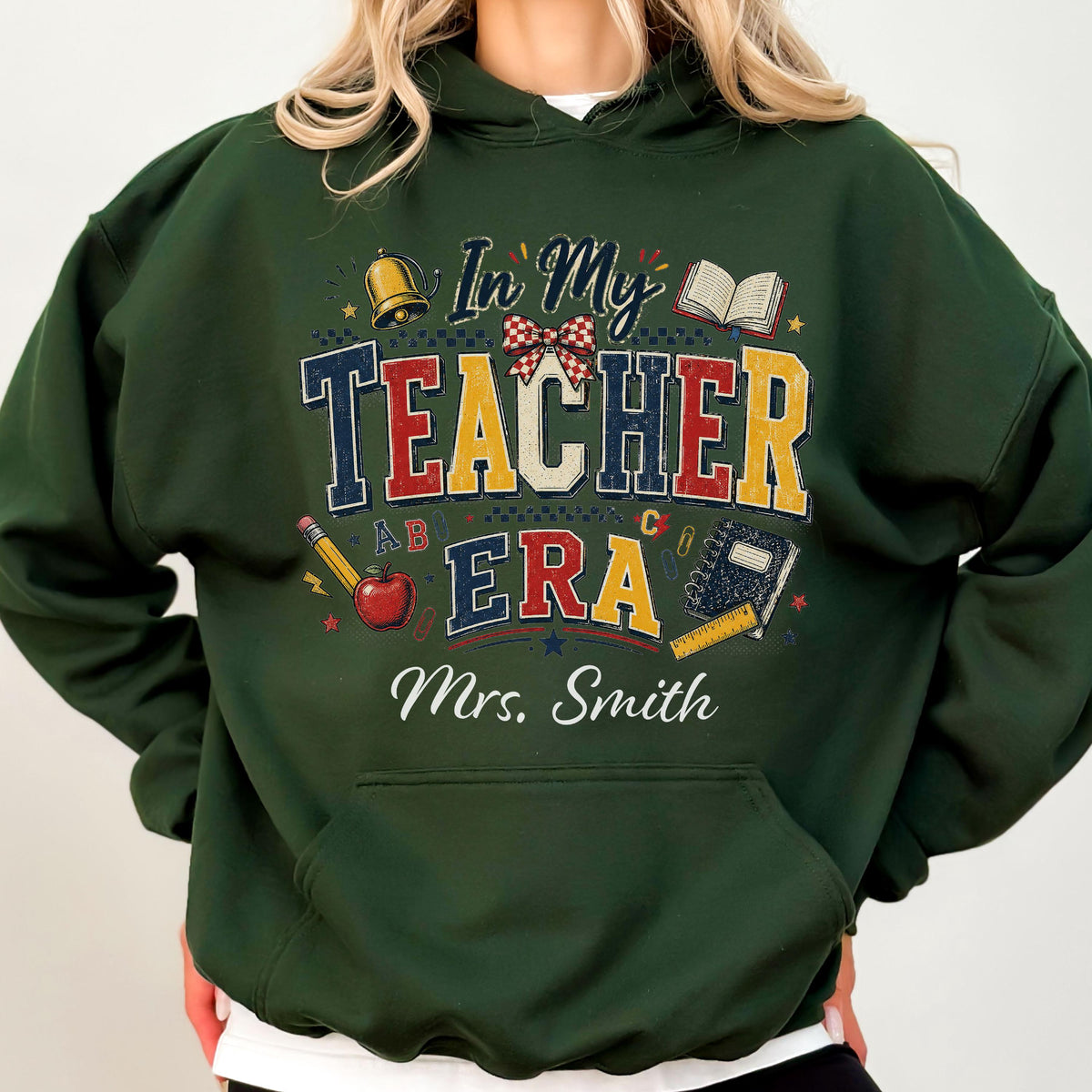 May include: A dark green hooded sweatshirt featuring a graphic print with the text In My Teacher Era and Mrs. Smith. The design includes illustrations of a gold bell, a red apple, a pencil, an open book, a notebook, and a red plaid bow.