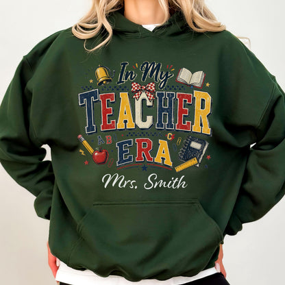 May include: A dark green hooded sweatshirt featuring a graphic print with the text In My Teacher Era and Mrs. Smith. The design includes illustrations of a gold bell, a red apple, a pencil, an open book, a notebook, and a red plaid bow.