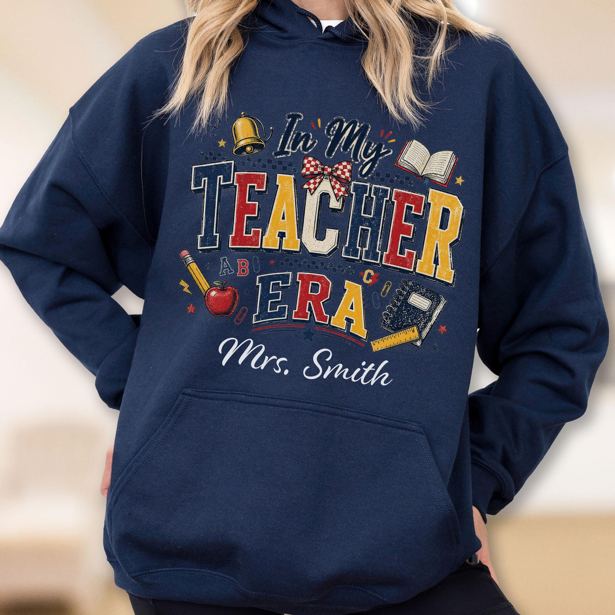 May include: A navy blue hooded sweatshirt featuring a graphic print with the text In My TEACHER ERA and Mrs. Smith. The design includes a gold bell, a red apple, a pencil, an open book, a notebook, a ruler, and a red and white checkered bow.