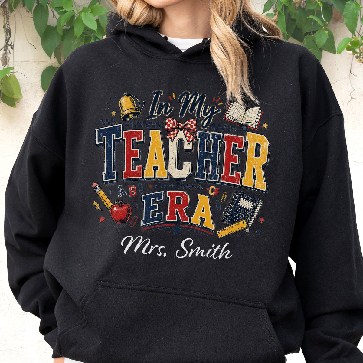 May include: A black hooded sweatshirt featuring a graphic print with the text In My Teacher Era and Mrs. Smith. The design includes a gold bell, an open book, a red apple with a pencil, a plaid bow, and a ruler, all in a distressed style.