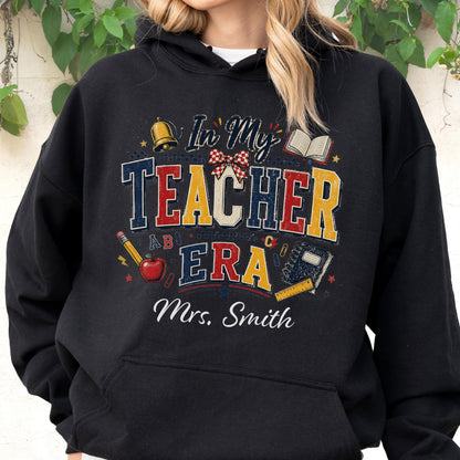 May include: A black hooded sweatshirt featuring a graphic print with the text In My Teacher Era and Mrs. Smith. The design includes a gold bell, an open book, a red apple with a pencil, a plaid bow, and a ruler, all in a distressed style.