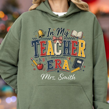 May include: An olive green hooded sweatshirt featuring a graphic print with the text In My Teacher Era and Mrs. Smith. The design includes a gold bell, an open book, a red apple, a pencil, a notebook, a ruler, and a red and white checkered bow.
