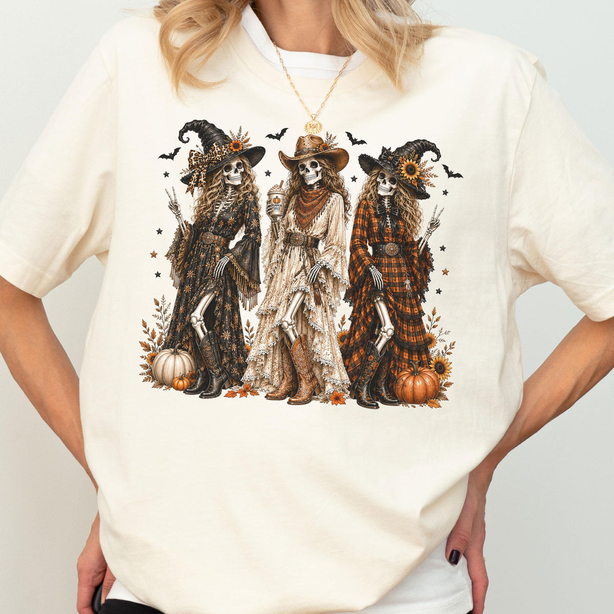 May include: A cream-colored t-shirt featuring a graphic of three skeletons dressed in western-style clothing, including hats, long dresses, and boots. The skeletons are surrounded by pumpkins, sunflowers, and small bats on a light background.