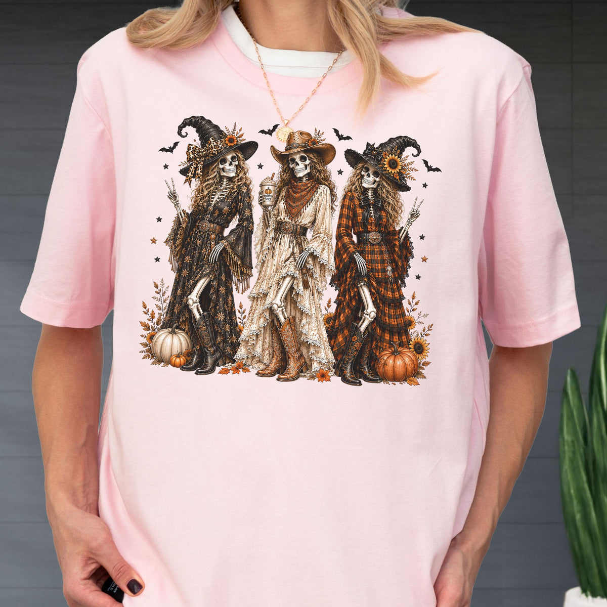 May include: A light pink t-shirt featuring a graphic print of three skeletons dressed in western-style clothing, including hats, long dresses, and boots. The skeletons are surrounded by pumpkins, sunflowers, and small bats on a light pink background.