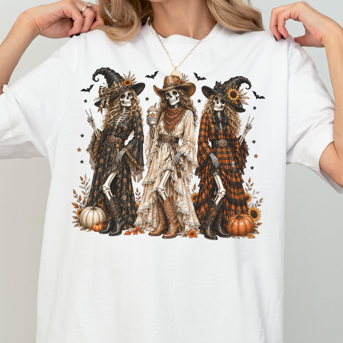 May include: A white t-shirt featuring a graphic of three skeletons dressed as cowgirls and witches. They wear hats, long dresses, and boots, surrounded by pumpkins, sunflowers, and bats. The central skeleton holds a drink, while the others pose.