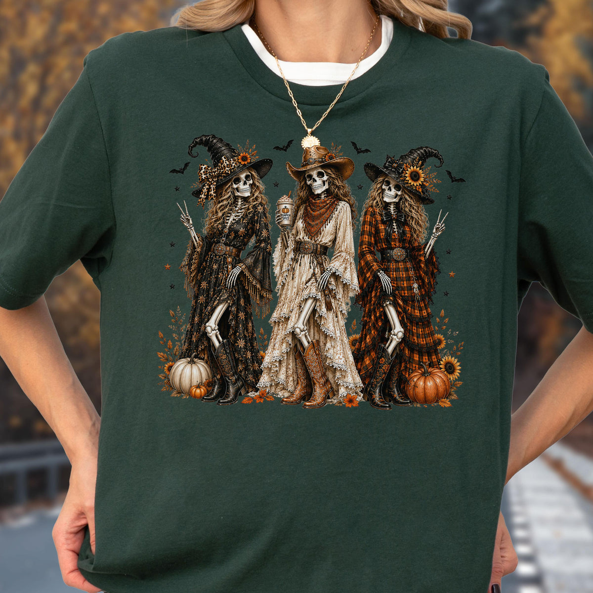May include: A dark green short-sleeve t-shirt featuring a graphic print of three skeletons dressed as witches in long dresses and cowboy boots. The skeletons are surrounded by pumpkins, sunflowers, and small bats. The center skeleton holds a cup.