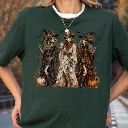 May include: A dark green short-sleeve t-shirt featuring a graphic print of three skeletons dressed as witches in long dresses and cowboy boots. The skeletons are surrounded by pumpkins, sunflowers, and small bats. The center skeleton holds a cup.