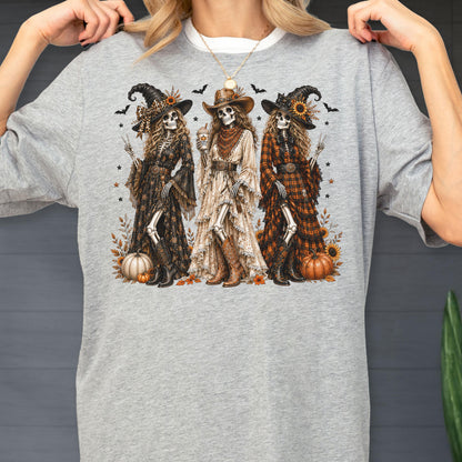 May include: A heather gray t-shirt featuring a graphic print of three skeletons dressed in western-style witch attire. The skeletons wear hats, long dresses, and boots, surrounded by pumpkins, sunflowers, and bats. The design includes autumn foliage.