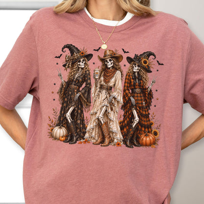 May include: A mauve short-sleeve t-shirt featuring a graphic print of three skeletons dressed in western-style witch attire, including hats, boots, and long dresses. The design includes sunflowers, pumpkins, bats, and stars surrounding the figures.