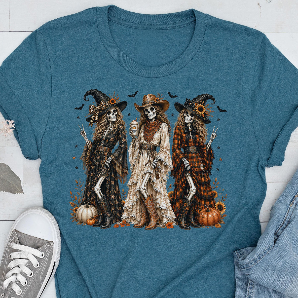 May include: A teal t-shirt featuring a graphic of three skeletons dressed in western-style clothing, including hats, long dresses, and boots. The skeletons are surrounded by pumpkins, sunflowers, and small bats, creating a detailed seasonal scene.