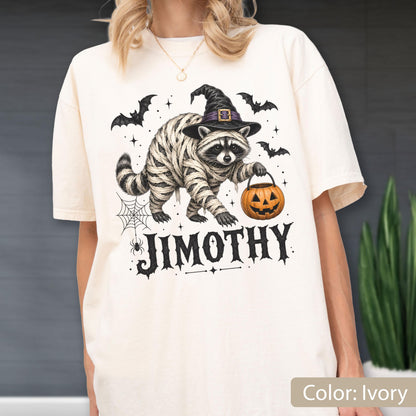 May include: An ivory t-shirt featuring a graphic of a raccoon wrapped in mummy bandages, wearing a black witch hat, and holding an orange jack-o'-lantern. The design includes black bats, a spider web, and the name JIMOTHY in bold black lettering.