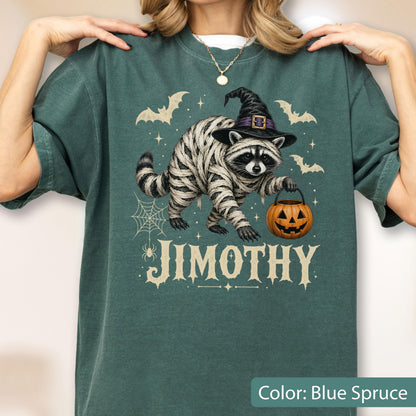 May include: A blue spruce t-shirt featuring a graphic of a raccoon wrapped in bandages like a mummy, wearing a black witch hat, and holding an orange jack-o'-lantern bucket. The design includes bats, a spider web, and the name JIMOTHY in white.