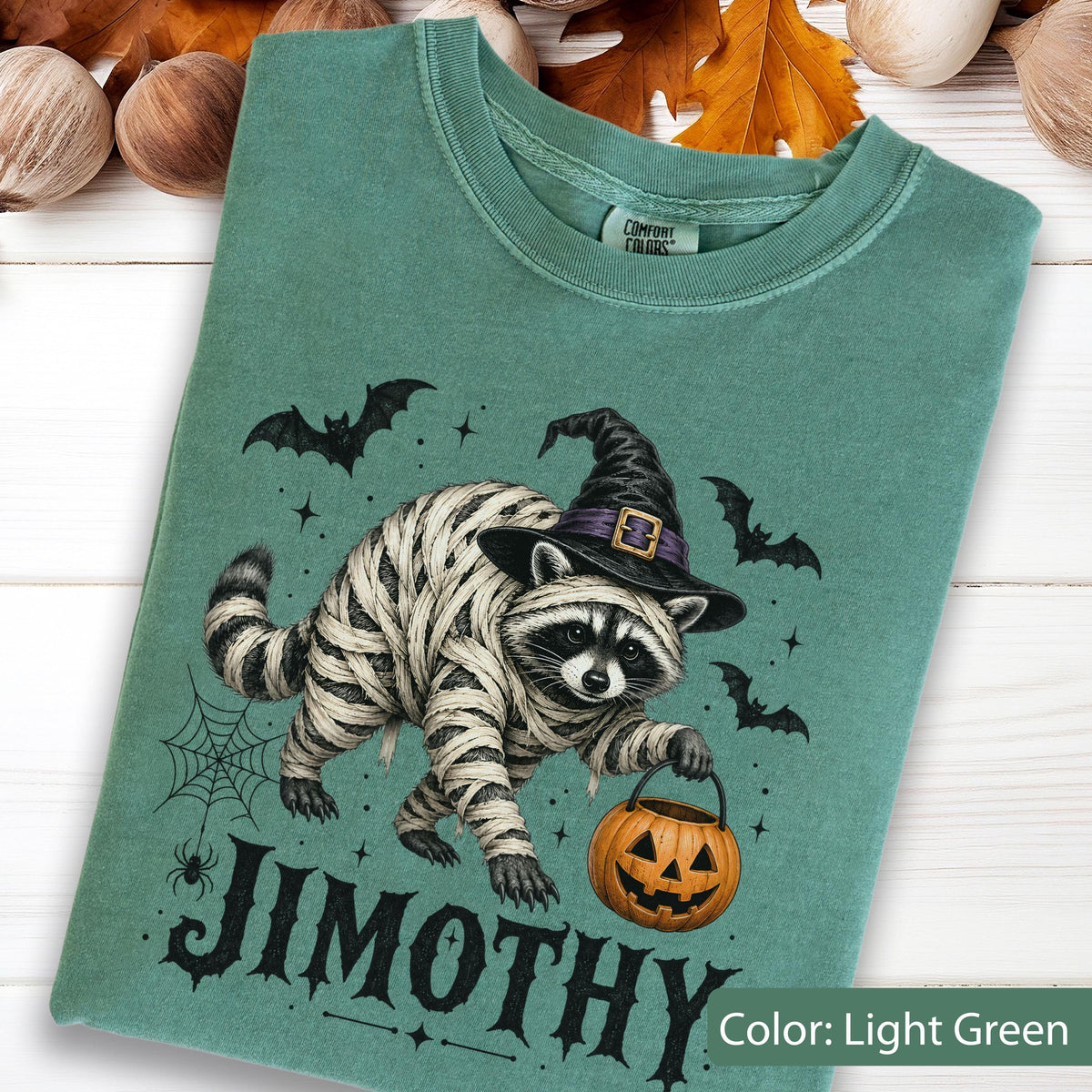 May include: A light green t-shirt featuring a graphic of a raccoon wrapped in white bandages like a mummy, wearing a black witch hat with a purple band. The raccoon holds an orange jack-o'-lantern bucket. The text JIMOTHY is printed below.