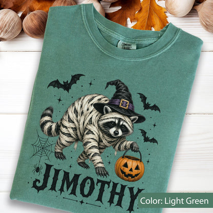 May include: A light green t-shirt featuring a graphic of a raccoon wrapped in white bandages like a mummy, wearing a black witch hat with a purple band. The raccoon holds an orange jack-o'-lantern bucket. The text JIMOTHY is printed below.