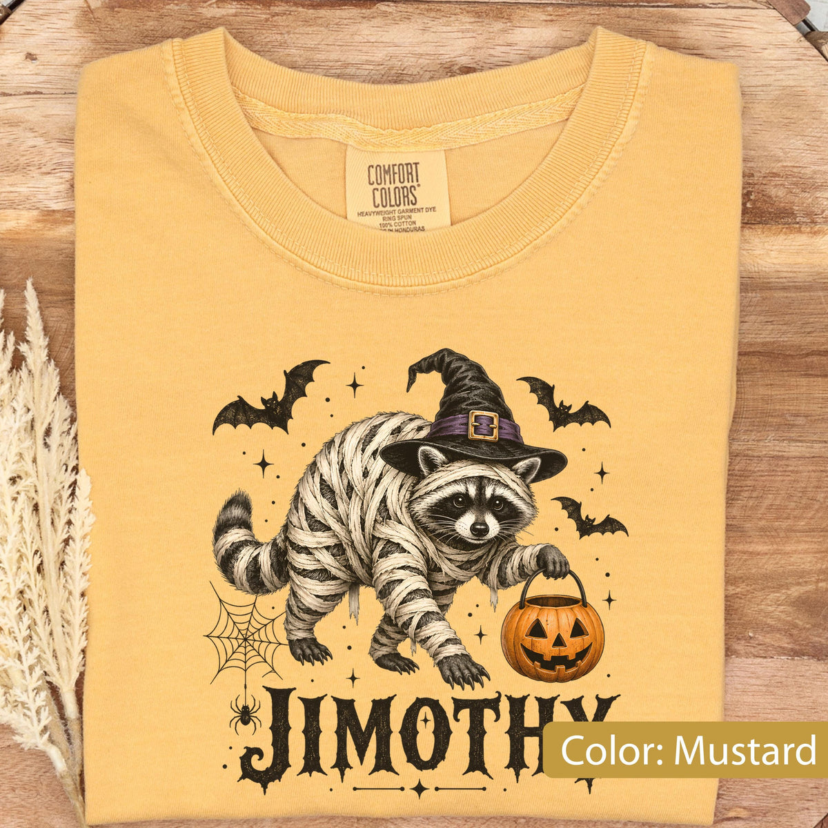 May include: A mustard yellow t-shirt featuring a graphic of a raccoon wrapped in white bandages like a mummy, wearing a black witch hat, and holding an orange jack-o'-lantern bucket. The design includes black bats, a spider web, and the text JIMOTHY.