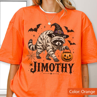 May include: An orange t-shirt featuring a graphic of a raccoon wrapped in white bandages, wearing a black witch hat, and holding an orange jack-o'-lantern. The design includes black bats, a spider web, and the text JIMOTHY in a bold black font.