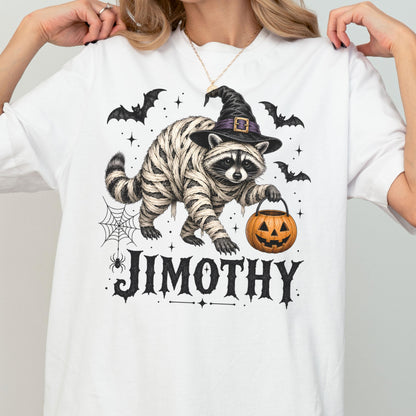 May include: A white t-shirt featuring a graphic of a raccoon wrapped in bandages like a mummy, wearing a black witch hat, and holding an orange jack-o'-lantern. The design includes black bats, a spider web, and the name JIMOTHY in black text.