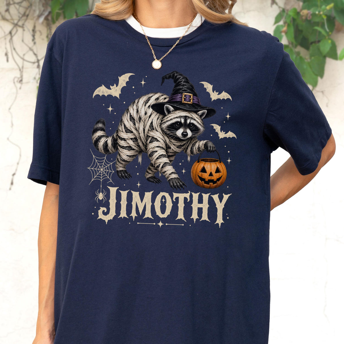 May include: A navy blue t-shirt featuring a graphic of a raccoon wrapped in white bandages like a mummy, wearing a black witch hat, and holding an orange jack-o'-lantern. The design includes bats, a spider web, and the text JIMOTHY in white.