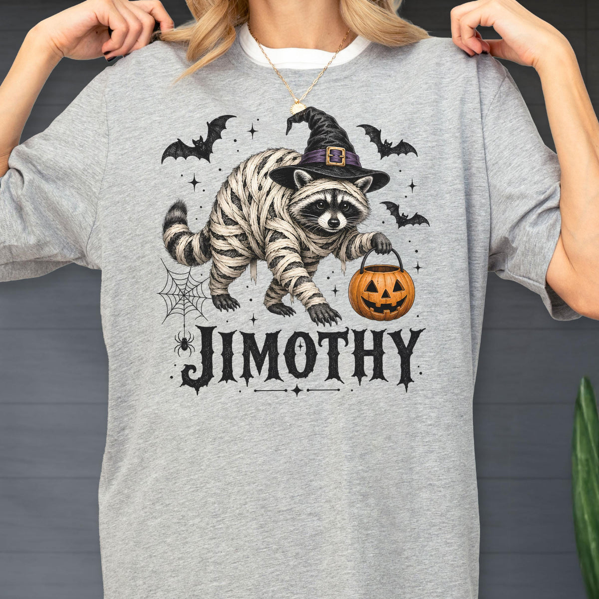 May include: A heather gray t-shirt featuring a graphic of a raccoon wrapped in mummy bandages, wearing a black witch hat, and holding an orange jack-o'-lantern. The design includes bats, a spider web, and the name JIMOTHY in black lettering.