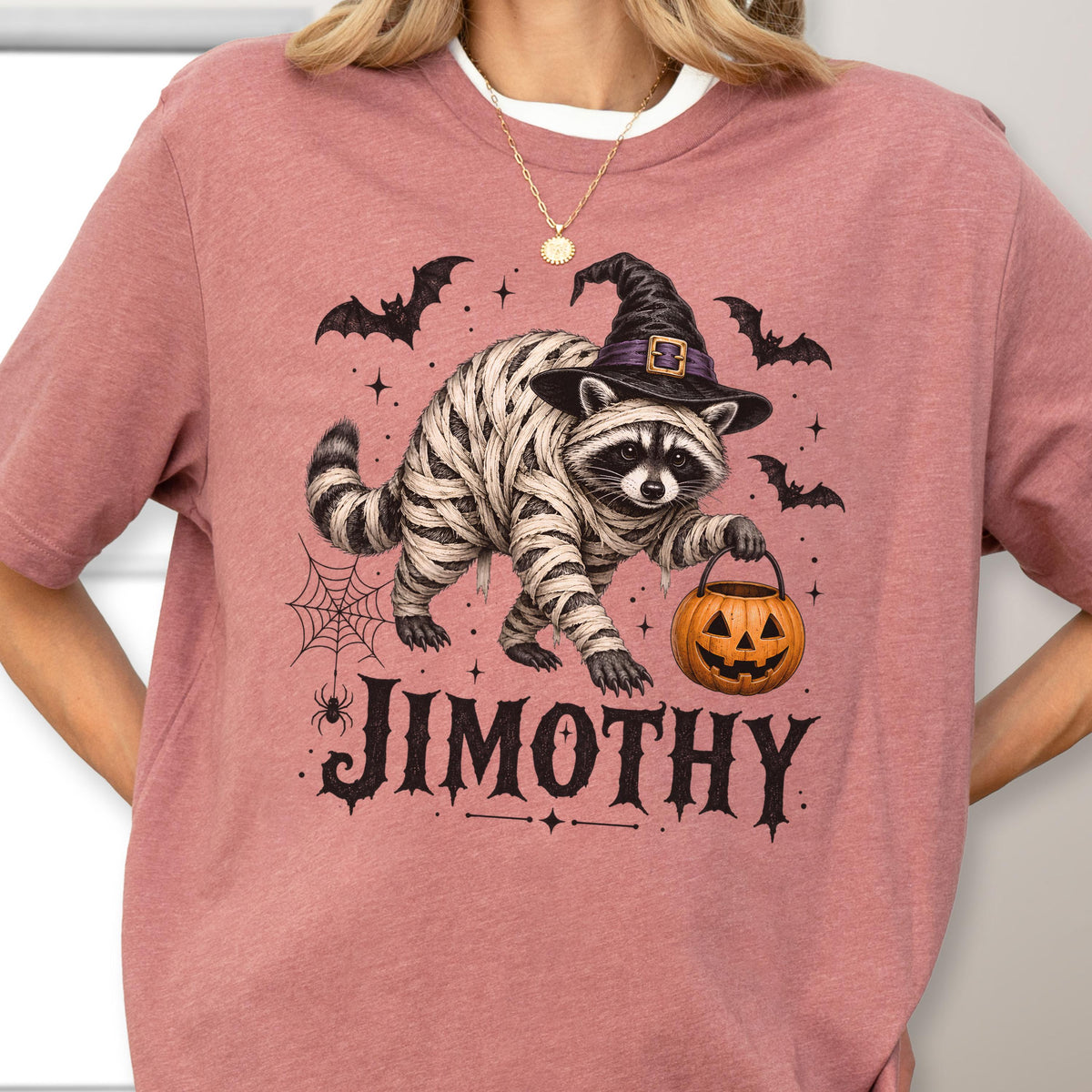 May include: A dusty rose t-shirt featuring a graphic of a raccoon wrapped in bandages like a mummy, wearing a black witch hat, and holding an orange jack-o'-lantern. The design includes bats, a spider web, and the name JIMOTHY in black text.