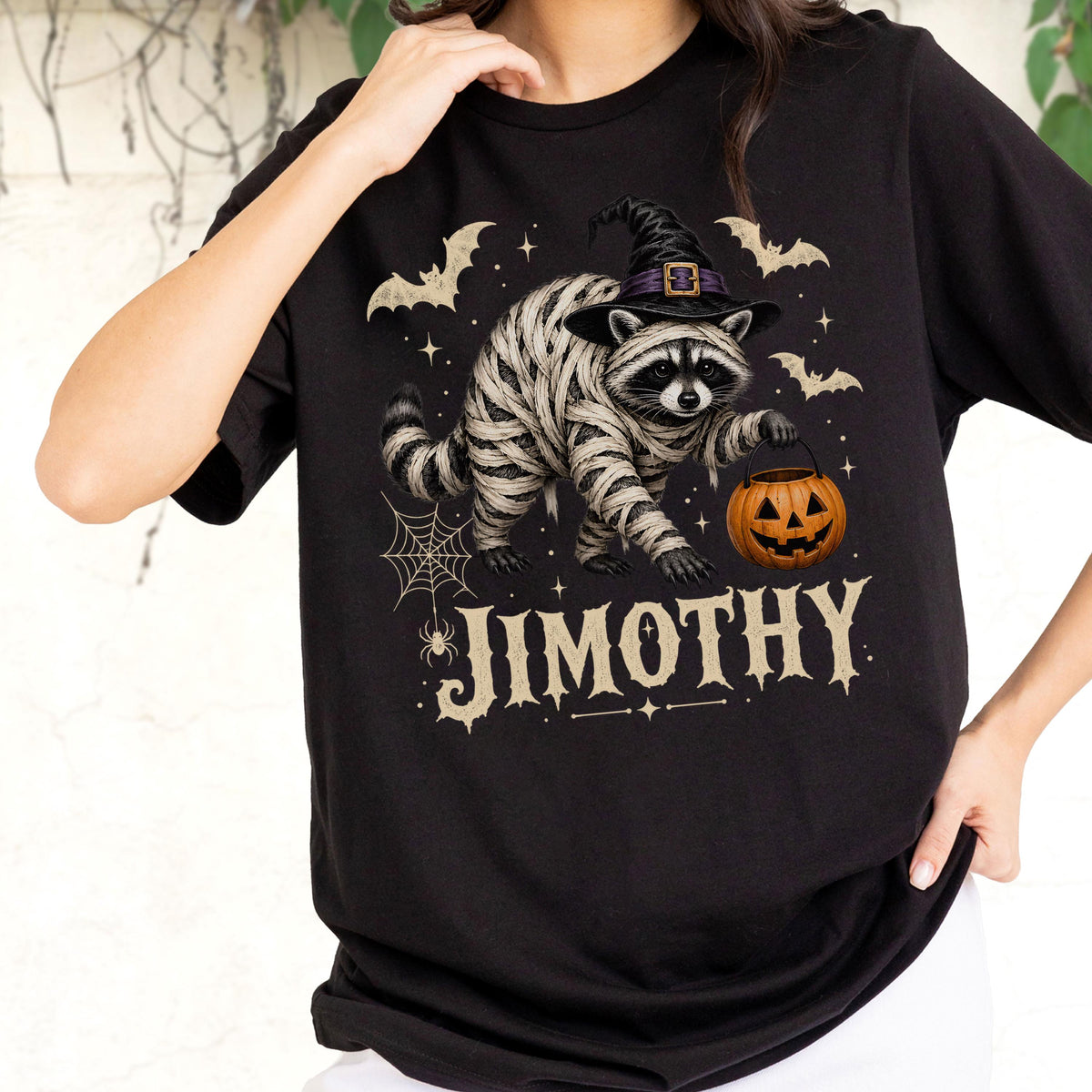 May include: A black short-sleeve t-shirt featuring a graphic of a raccoon wrapped in bandages like a mummy, wearing a black witch hat, holding an orange jack-o'-lantern, surrounded by bats, a spider web, and the text JIMOTHY in a beige font.