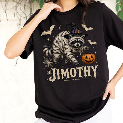 May include: A black short-sleeve t-shirt featuring a graphic of a raccoon wrapped in bandages like a mummy, wearing a black witch hat, holding an orange jack-o'-lantern, surrounded by bats, a spider web, and the text JIMOTHY in a beige font.
