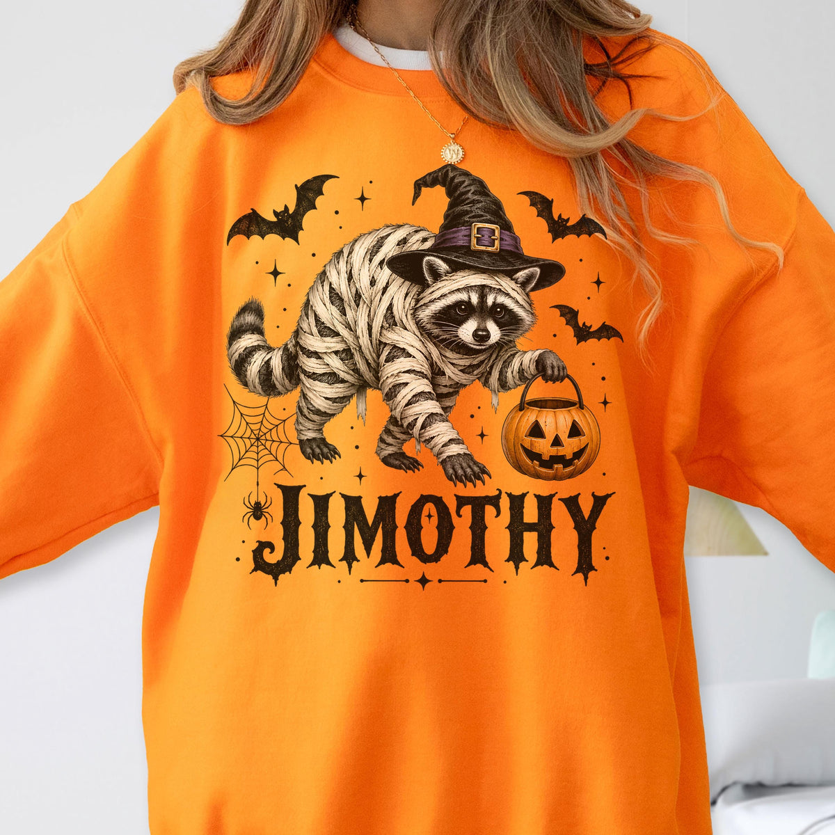May include: A bright orange sweatshirt featuring a graphic of a raccoon wrapped in white bandages like a mummy, wearing a black witch hat, and holding a jack-o'-lantern. The design includes bats, a spider web, and the text JIMOTHY in black.