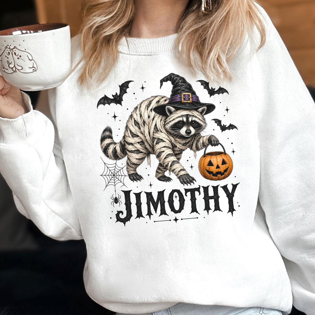 May include: A white long-sleeve sweatshirt featuring a graphic of a raccoon dressed as a mummy wearing a witch hat and holding a jack-o'-lantern. The design includes bats, a spider web, and the name JIMOTHY printed in a bold black font.