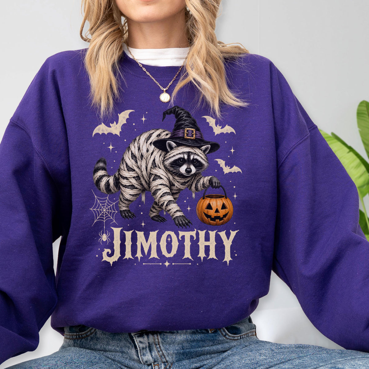 May include: A purple long-sleeve sweatshirt featuring a graphic of a raccoon wrapped in bandages, wearing a black witch hat, and holding an orange jack-o'-lantern. The design includes bats, a spider web, and the text JIMOTHY in white lettering.