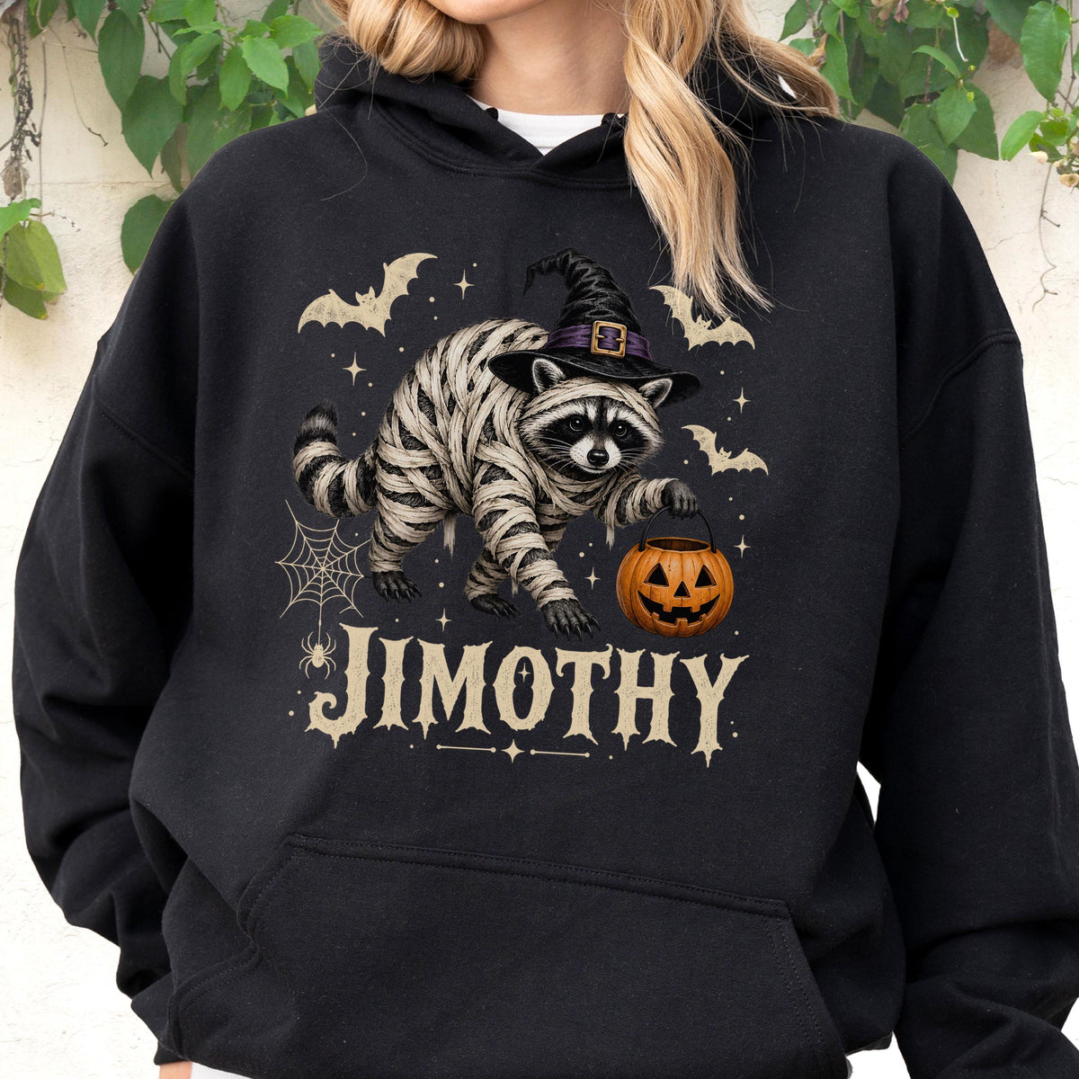 May include: A black hooded sweatshirt featuring a graphic of a raccoon wrapped in bandages like a mummy, wearing a black witch hat. The raccoon holds an orange jack-o'-lantern bucket. Bats, a spider web, and the name JIMOTHY are printed below.