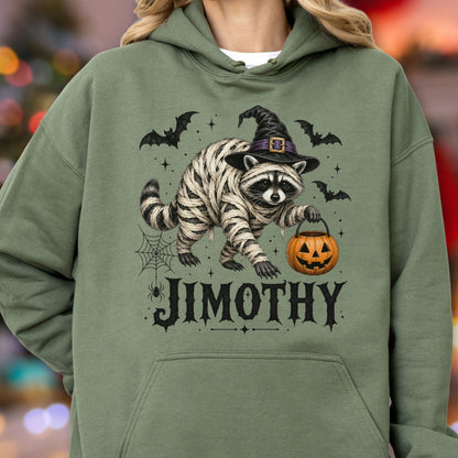 May include: An olive green hooded sweatshirt featuring a graphic of a raccoon wrapped in white bandages, wearing a black witch hat, and holding an orange jack-o'-lantern bucket. The design includes black bats, a spider web, and the name JIMOTHY.