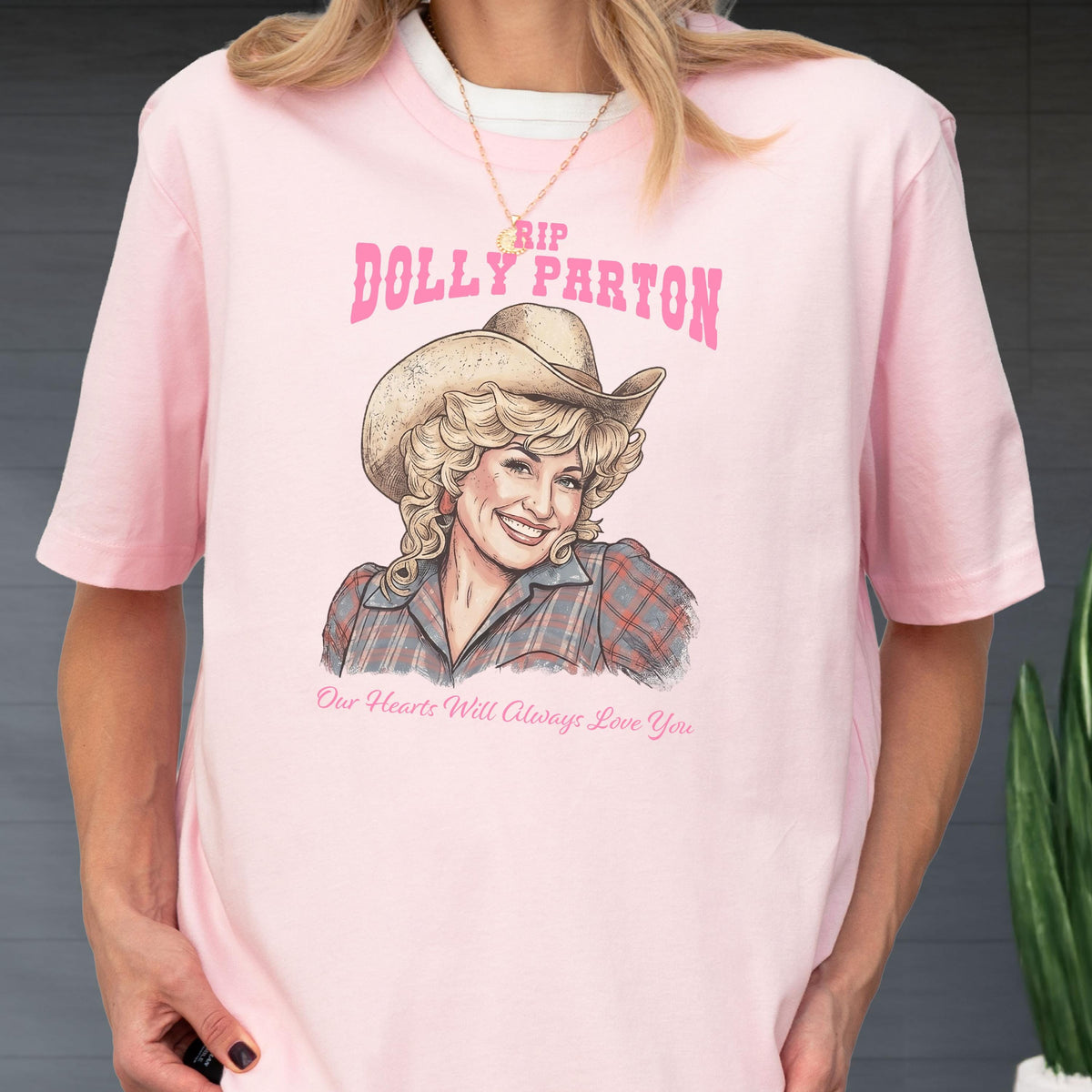 May include: A light pink t-shirt featuring a graphic illustration of Dolly Parton wearing a cowboy hat and a plaid shirt. The text above reads RIP DOLLY PARTON in pink, and the text below reads Our Hearts Will Always Love You in pink.