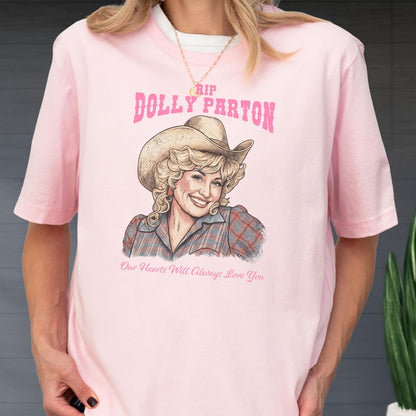 May include: A light pink t-shirt featuring a graphic illustration of Dolly Parton wearing a cowboy hat and a plaid shirt. The text above reads RIP DOLLY PARTON in pink, and the text below reads Our Hearts Will Always Love You in pink.