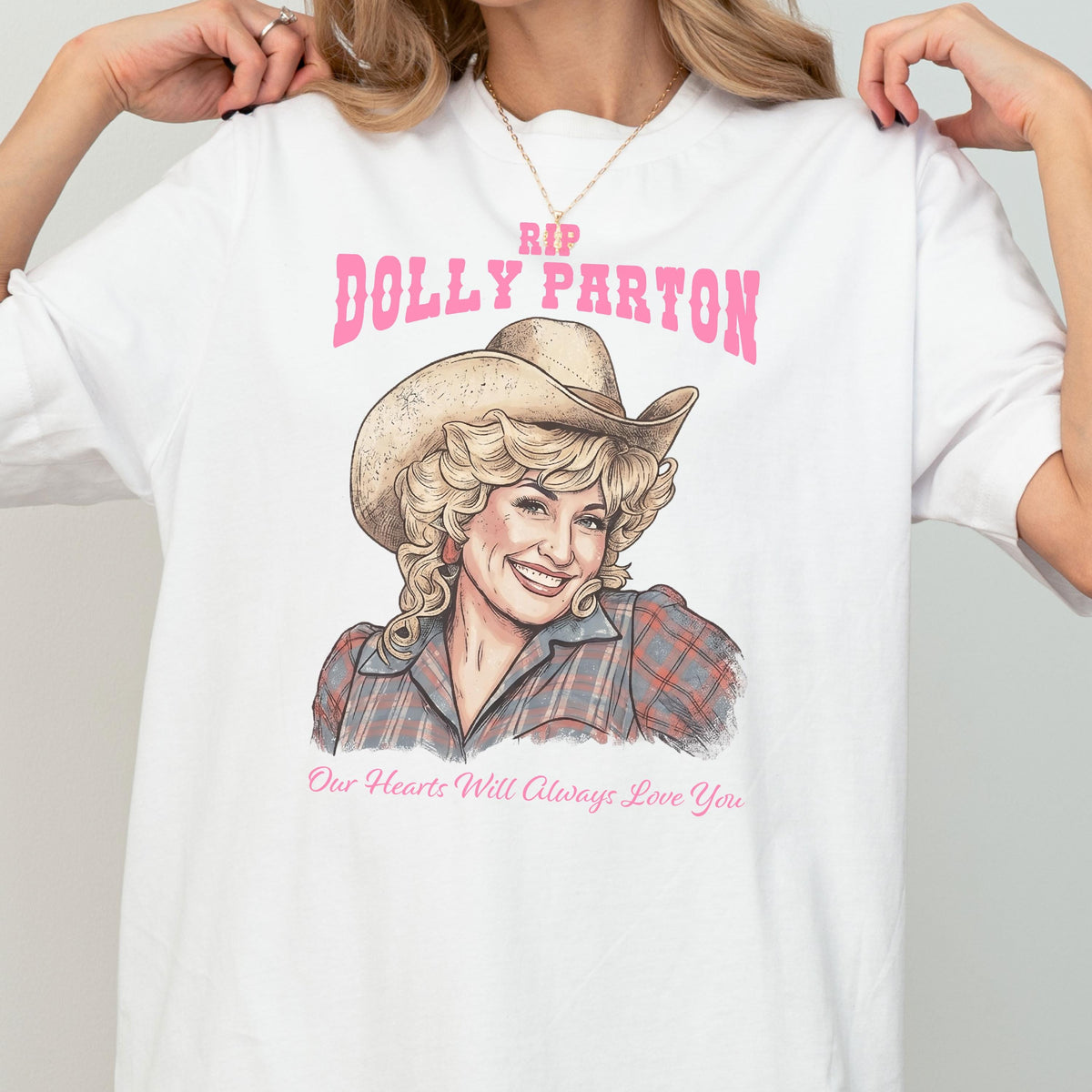 May include: A white t-shirt featuring a graphic illustration of Dolly Parton wearing a cowboy hat and a plaid shirt. The text above reads RIP DOLLY PARTON in pink, and the text below reads Our Hearts Will Always Love You in pink script.
