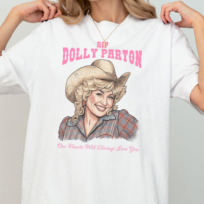 May include: A white t-shirt featuring a graphic illustration of Dolly Parton wearing a cowboy hat and a plaid shirt. The text above reads RIP DOLLY PARTON in pink, and the text below reads Our Hearts Will Always Love You in pink script.