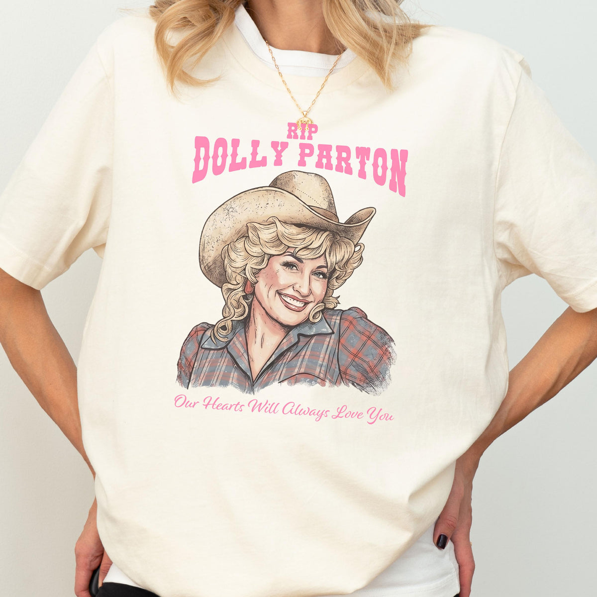 May include: A cream-colored t-shirt featuring a graphic illustration of Dolly Parton wearing a cowboy hat and a plaid shirt. The text reads RIP DOLLY PARTON in pink letters above the illustration and Our Hearts Will Always Love You below.