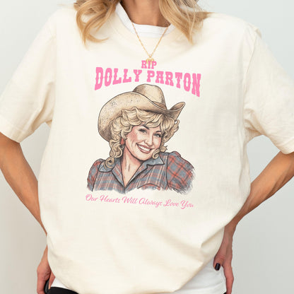 May include: A cream-colored t-shirt featuring a graphic illustration of Dolly Parton wearing a cowboy hat and a plaid shirt. The text reads RIP DOLLY PARTON in pink letters above the illustration and Our Hearts Will Always Love You below.