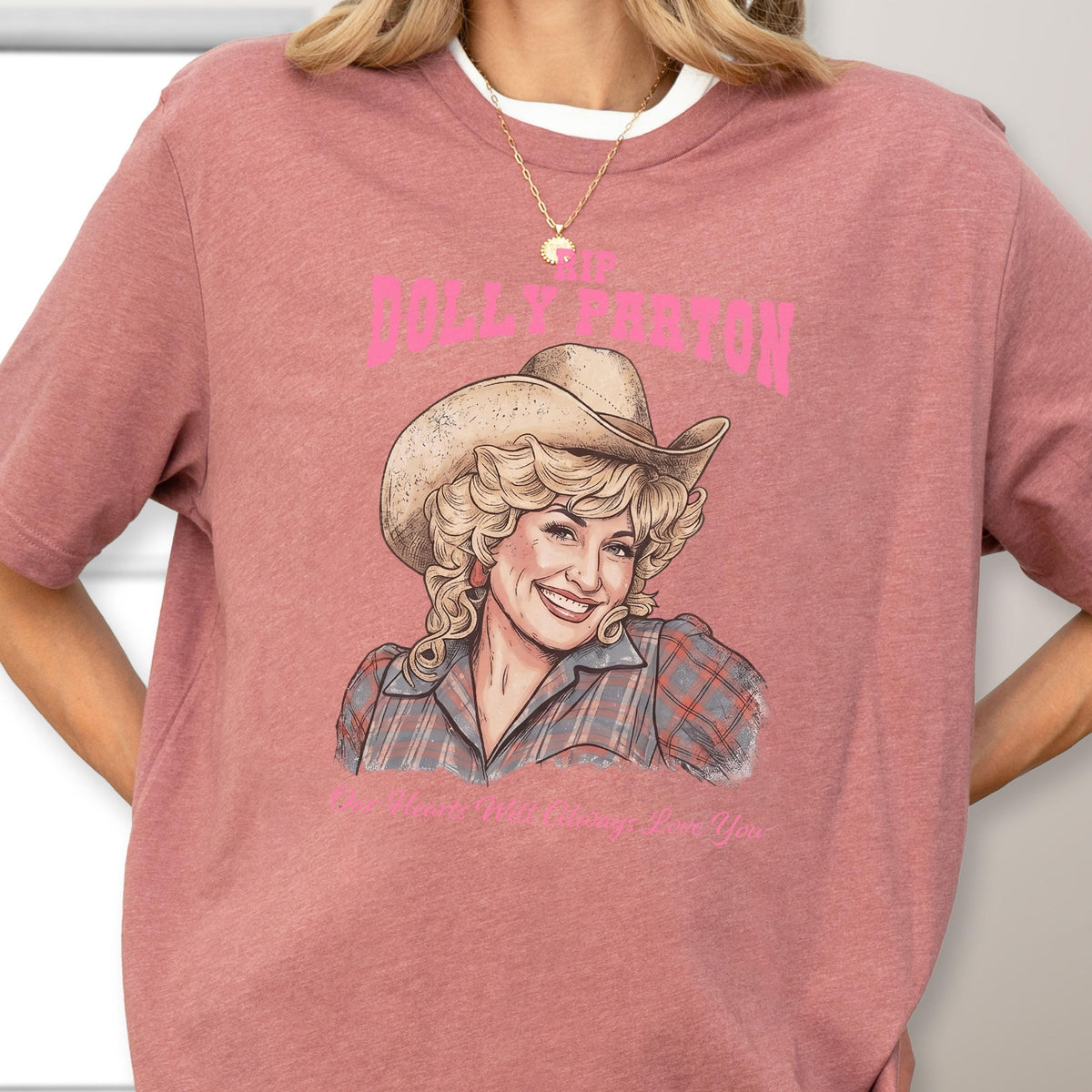 May include: A dusty rose short-sleeve t-shirt featuring a graphic illustration of Dolly Parton wearing a cowboy hat and a plaid shirt. The text "RIP DOLLY PARTON" appears in pink above the illustration, with additional text below the image.