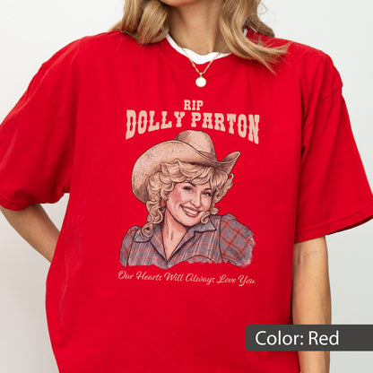 May include: A bright red short-sleeved t-shirt featuring a graphic illustration of Dolly Parton wearing a cowboy hat and a plaid shirt. The text reads RIP DOLLY PARTON above the illustration and Our Hearts Will Always Love You below it.