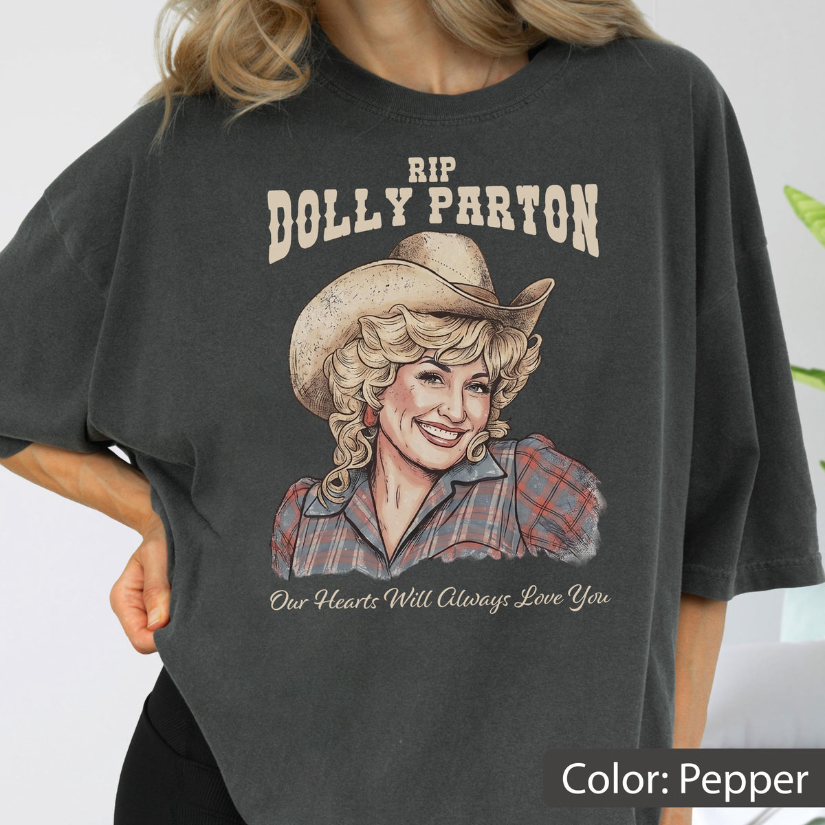 May include: A charcoal gray t-shirt featuring a graphic illustration of Dolly Parton wearing a cowboy hat and a plaid shirt. The text above reads RIP DOLLY PARTON and the text below reads Our Hearts Will Always Love You in a script font.