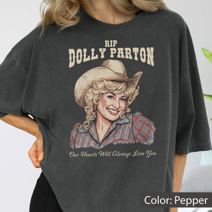 May include: A charcoal gray t-shirt featuring a graphic illustration of Dolly Parton wearing a cowboy hat and a plaid shirt. The text above reads RIP DOLLY PARTON and the text below reads Our Hearts Will Always Love You in a script font.