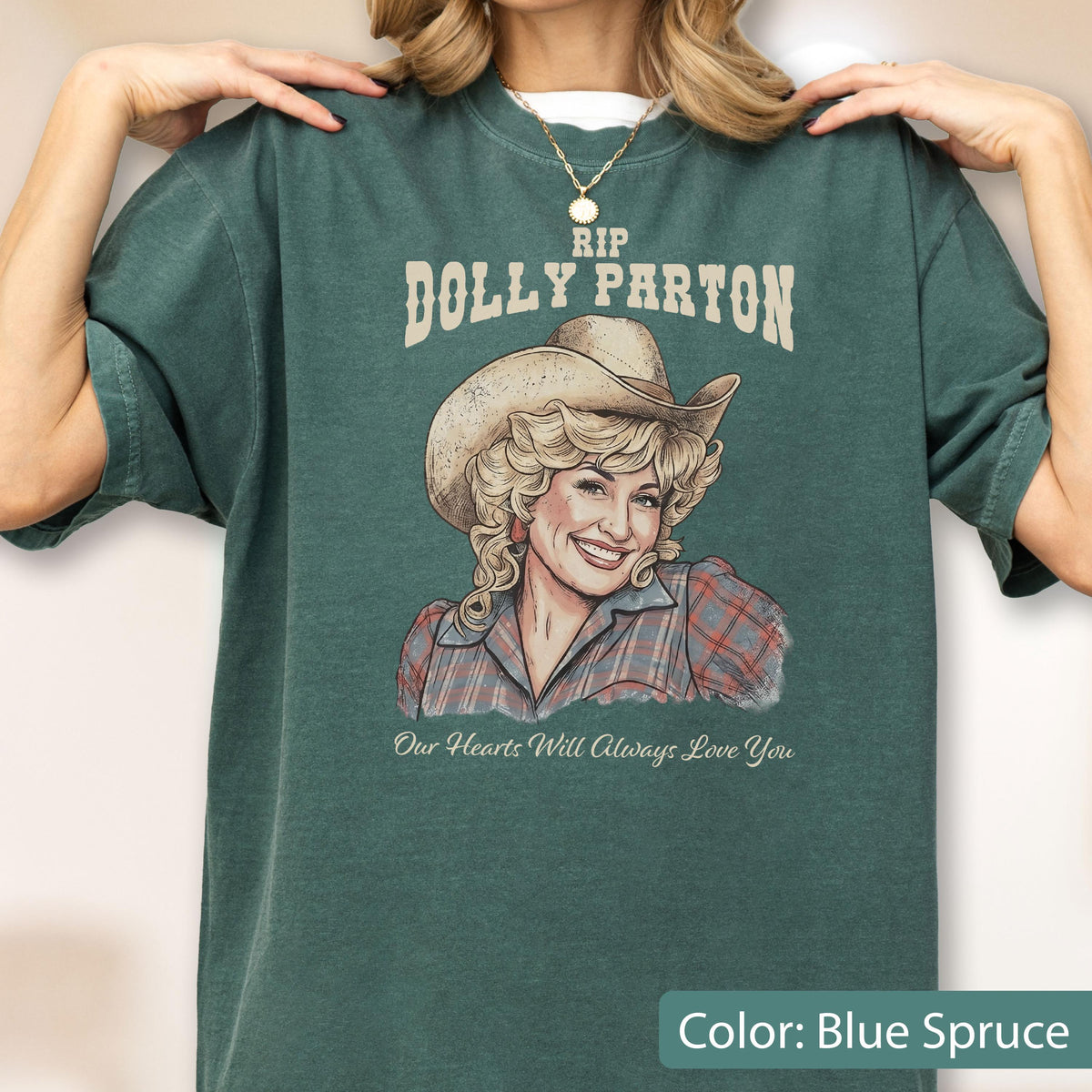 May include: A blue spruce t-shirt featuring a graphic illustration of Dolly Parton wearing a cowboy hat and a plaid shirt. The text reads RIP DOLLY PARTON above the illustration and Our Hearts Will Always Love You below the illustration.