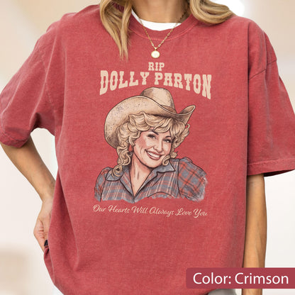 May include: A crimson short-sleeve t-shirt featuring a graphic illustration of Dolly Parton wearing a cowboy hat. The text reads RIP DOLLY PARTON and Our Hearts Will Always Love You. The shirt has a crew neckline and a relaxed, boxy fit.